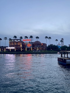 A waterfront view during twilight features a large entertainment or restaurant building with neon signs, surrounded by tall palm trees. The reflection of the building's lights shimmers on the calm water. A boat with an American flag is moored in the lower right corner.