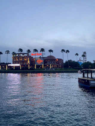 A waterfront view during twilight features a large entertainment or restaurant building with neon signs, surrounded by tall palm trees. The reflection of the building's lights shimmers on the calm water. A boat with an American flag is moored in the lower right corner.