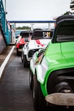 A lineup of colorful go-karts is positioned on a racetrack. The go-karts are brightly colored in green, white, and red, with distinct black seats and visible roll bars. The setting appears to be a motorsports facility, with barriers and spectators seen in the background.