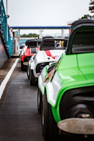 A lineup of colorful go-karts is positioned on a racetrack. The go-karts are brightly colored in green, white, and red, with distinct black seats and visible roll bars. The setting appears to be a motorsports facility, with barriers and spectators seen in the background.