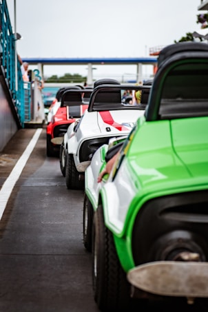 A lineup of colorful go-karts is positioned on a racetrack. The go-karts are brightly colored in green, white, and red, with distinct black seats and visible roll bars. The setting appears to be a motorsports facility, with barriers and spectators seen in the background.