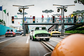 A colorful amusement park setting with a track for small cars designed for children or families. Several cars of different colors, including green, yellow, red, and blue, are aligned on the track. There are people walking on an overhead pedestrian bridge, observing the activity below. The environment includes traffic lights and decorative elements.