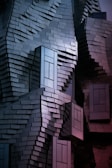 A building facade made of uniquely arranged rectangular metal panels creating an abstract and textured appearance. The panels reflect light in a way that produces a gradient of colors, and the structure has protruding window-like sections.