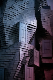 A building facade made of uniquely arranged rectangular metal panels creating an abstract and textured appearance. The panels reflect light in a way that produces a gradient of colors, and the structure has protruding window-like sections.
