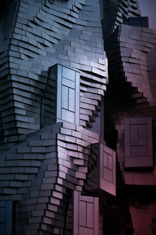 A building facade made of uniquely arranged rectangular metal panels creating an abstract and textured appearance. The panels reflect light in a way that produces a gradient of colors, and the structure has protruding window-like sections.