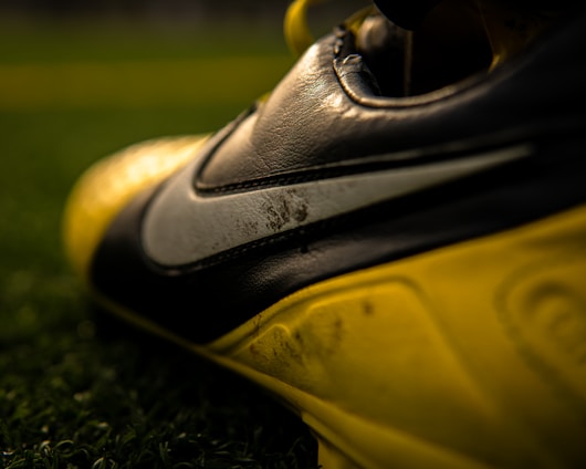 A close-up view of a football or soccer shoe with a distinctive swoosh logo in focus, featuring scuffed and worn textures on the leather surface. The shoe is predominantly black and yellow, with rich detailing captured under gleaming light. The background includes blurred green artificial turf, suggesting a sports field setting.