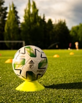 A soccer ball with green and white patterns is balanced on a yellow cone, placed on a lush green field under sunlight. Orange cones are positioned in the background, with trees bordering the field.