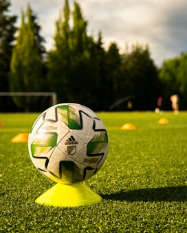 A soccer ball with green and white patterns is balanced on a yellow cone, placed on a lush green field under sunlight. Orange cones are positioned in the background, with trees bordering the field.