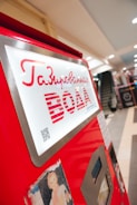 A bright red vending machine with Russian text offering drinks, placed in a spacious indoor area with a modern design. The machine has a metallic frame and a small screen, with visible buttons and a QR code.
