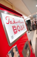 A bright red vending machine with Russian text offering drinks, placed in a spacious indoor area with a modern design. The machine has a metallic frame and a small screen, with visible buttons and a QR code.