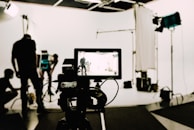 In a professional studio setting, a camera is focused on several people working in the background with various lighting and film equipment. The camera display shows a mirrored view of the scene being shot, while crew members adjust lights and arrange equipment.