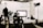 In a professional studio setting, a camera is focused on several people working in the background with various lighting and film equipment. The camera display shows a mirrored view of the scene being shot, while crew members adjust lights and arrange equipment.