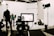 In a professional studio setting, a camera is focused on several people working in the background with various lighting and film equipment. The camera display shows a mirrored view of the scene being shot, while crew members adjust lights and arrange equipment.