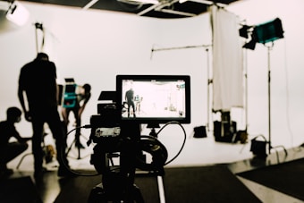 In a professional studio setting, a camera is focused on several people working in the background with various lighting and film equipment. The camera display shows a mirrored view of the scene being shot, while crew members adjust lights and arrange equipment.
