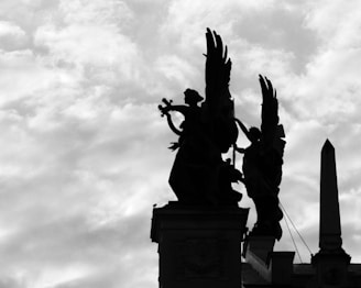 Silhouettes of statues with winged figures stand prominently against a cloudy sky. One figure appears to be playing a musical instrument while the other leans forward, creating a dramatic and solemn scene.