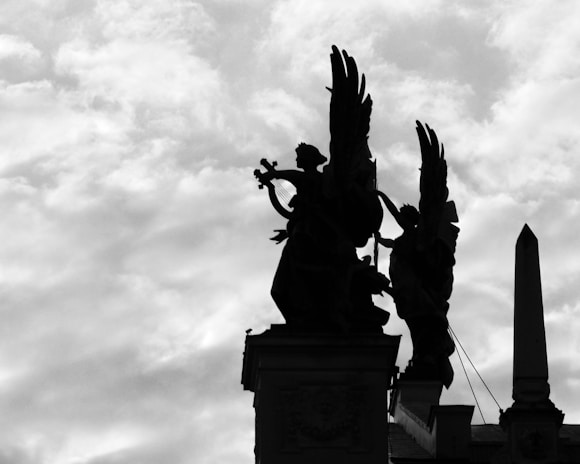 Silhouettes of statues with winged figures stand prominently against a cloudy sky. One figure appears to be playing a musical instrument while the other leans forward, creating a dramatic and solemn scene.