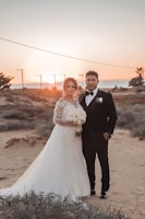 A couple stands on a sandy landscape during sunset. The woman is wearing a white wedding gown, holding a bouquet of white flowers, while the man is in a black tuxedo with a white shirt and bow tie. Sparse vegetation and the ocean can be seen in the background, with a clear sky transitioning from orange to blue.