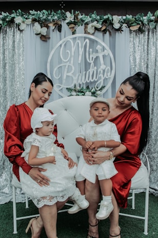 Two women in matching red dresses hold two toddlers, each dressed in white, sitting on a white chair. The backdrop features a decorative floral arrangement with the words 'Mi Bautizo' in the center.