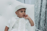 Stylish toddler in a cream-colored occasion wear outfit, standing by a rustic wooden chair.