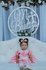 A young child wearing a pink dress and Minnie Mouse ears sits on a white tufted sofa. Behind the child is a circular sign with the text 'Mi Bautizo' adorned with flowers and foliage. The background consists of sheer, draped fabric and additional white and green floral arrangements above.