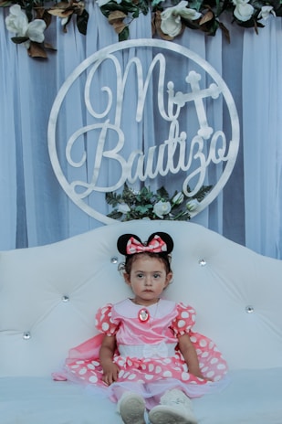 A young child wearing a pink dress and Minnie Mouse ears sits on a white tufted sofa. Behind the child is a circular sign with the text 'Mi Bautizo' adorned with flowers and foliage. The background consists of sheer, draped fabric and additional white and green floral arrangements above.