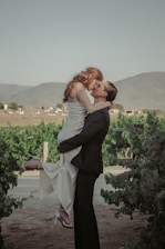 A couple sharing a kiss, with the man lifting the woman up in an affectionate embrace. They are surrounded by lush green vineyards, with rolling hills in the background under a clear sky. The woman is wearing a white dress and heels, while the man is dressed in a dark suit.