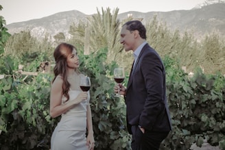 A woman in a white dress and a man in a dark suit are standing together, holding glasses of red wine, surrounded by lush green grapevines with mountains in the background.