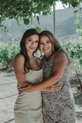 Two smiling women are embracing under a lush green canopy, possibly in a vineyard with hills in the background. One is wearing a white dress and the other a patterned dress. The setting has a serene and natural vibe.