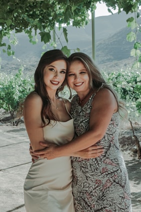 Two smiling women are embracing under a lush green canopy, possibly in a vineyard with hills in the background. One is wearing a white dress and the other a patterned dress. The setting has a serene and natural vibe.