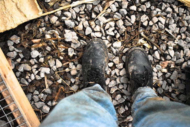 A pair of worn black boots standing on a surface covered with small gray gravel and twigs. The lower part of blue jeans is visible, suggesting a casual or work setting. Surrounding elements include pieces of wood and wire mesh in the corners.