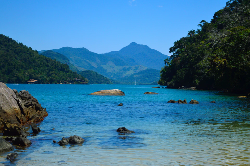 The image depicts a serene tropical beach scene with a large rock formation on the left side, surrounded by clear blue water and lush green trees. The sky above is a bright blue color, suggesting a sunny day. In the distance, there are mountains rising against the horizon, adding depth to the landscape.