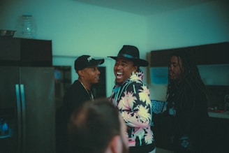 Three people are gathered in a dimly lit room, engaged in a lively conversation. One person in the middle is laughing joyfully, wearing a hat and a colorful, patterned hoodie, while the others look on with smiles.