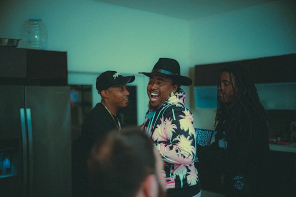 Three people are gathered in a dimly lit room, engaged in a lively conversation. One person in the middle is laughing joyfully, wearing a hat and a colorful, patterned hoodie, while the others look on with smiles.