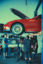 A car is elevated on a lift in an automotive garage, with its hood open. Three individuals are gathered around the car, seemingly involved in a discussion or working together. Various tools and equipment are visible in the background, indicating a workspace dedicated to vehicle maintenance or repair.