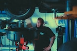 A mechanic stands in a garage near a lifted car tire, surrounded by various automotive tools and equipment. He has a beard, tattoos on his forearm, and is wearing a dark t-shirt with a logo. The background includes shelves with items and parts, and a blue and white wall sign.