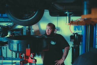 A mechanic stands in a garage near a lifted car tire, surrounded by various automotive tools and equipment. He has a beard, tattoos on his forearm, and is wearing a dark t-shirt with a logo. The background includes shelves with items and parts, and a blue and white wall sign.