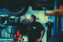 A mechanic stands in a garage near a lifted car tire, surrounded by various automotive tools and equipment. He has a beard, tattoos on his forearm, and is wearing a dark t-shirt with a logo. The background includes shelves with items and parts, and a blue and white wall sign.