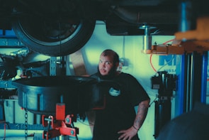 A mechanic stands in a garage near a lifted car tire, surrounded by various automotive tools and equipment. He has a beard, tattoos on his forearm, and is wearing a dark t-shirt with a logo. The background includes shelves with items and parts, and a blue and white wall sign.
