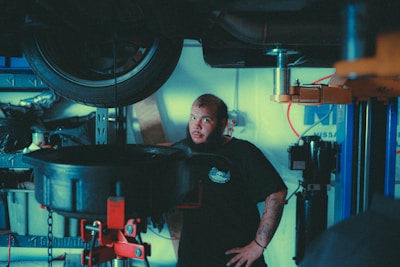 A mechanic stands in a garage near a lifted car tire, surrounded by various automotive tools and equipment. He has a beard, tattoos on his forearm, and is wearing a dark t-shirt with a logo. The background includes shelves with items and parts, and a blue and white wall sign.
