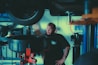 A mechanic stands in a garage near a lifted car tire, surrounded by various automotive tools and equipment. He has a beard, tattoos on his forearm, and is wearing a dark t-shirt with a logo. The background includes shelves with items and parts, and a blue and white wall sign.