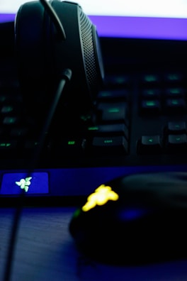 A close-up of a gaming setup, featuring a headset placed on a keyboard and a gaming mouse with an illuminated emblem. The lighting is dim, with a strong emphasis on the neon blue and yellow accents on the devices.