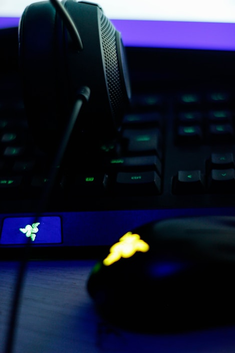 A close-up of a gaming setup, featuring a headset placed on a keyboard and a gaming mouse with an illuminated emblem. The lighting is dim, with a strong emphasis on the neon blue and yellow accents on the devices.