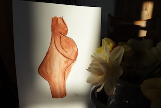 A watercolor painting on paper showing an abstract form with a curvy shape, primarily in shades of brown. The painting is on a white piece of paper, standing on a dark surface. Beside the paper is a ceramic vase containing yellow and white flowers illuminated by a soft light.