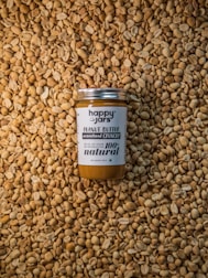 A jar of peanut butter labeled 'Happy Jars' and '100% natural' is centered on a large pile of unshelled peanuts. The design of the jar emphasizes its natural and preservative-free qualities.