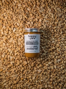 A jar of peanut butter labeled 'Happy Jars' and '100% natural' is centered on a large pile of unshelled peanuts. The design of the jar emphasizes its natural and preservative-free qualities.