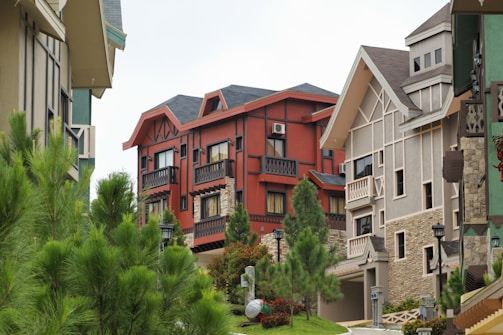 A picturesque residential area features a row of modern, multistory houses with varied architectural designs. The houses are adorned with stone facades, wooden details, and different colored walls. Lush green pine trees and well-maintained shrubs line the street, contributing to a serene suburban atmosphere.
