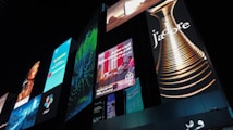 A series of illuminated advertisements displayed on a large, modern building at night. The ads include a variety of images and text, such as a perfume bottle with the word 'J'adore', a woman's portrait, and several panels with colorful designs and text in different languages.