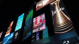 A series of illuminated advertisements displayed on a large, modern building at night. The ads include a variety of images and text, such as a perfume bottle with the word 'J'adore', a woman's portrait, and several panels with colorful designs and text in different languages.