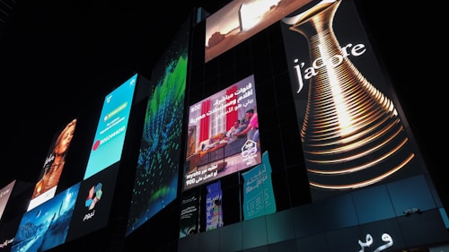 A series of illuminated advertisements displayed on a large, modern building at night. The ads include a variety of images and text, such as a perfume bottle with the word 'J'adore', a woman's portrait, and several panels with colorful designs and text in different languages.