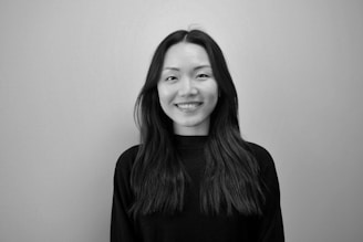 A black and white portrait of a person with long, dark hair wearing a dark top. The person is smiling and looking directly at the camera, set against a plain background.